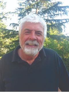 A white man with copious smile wrinkles and white hair and a beard sitting in front of a tree