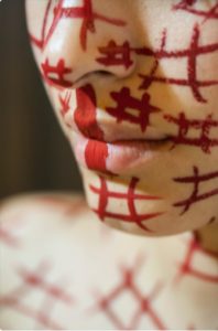 A face covered in white makeup with red hashtags drawn on