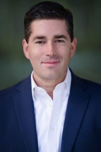 Gregory Phillips with short brown hair wearing a suit with no tie. 