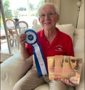 A white man in a red polo holdingup a Grand Prize Ribbon and The Devil's Bookkeeper Trilogy