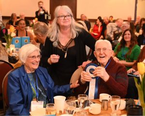 100 year-old Burl Harmon sits at a table with his wife while receiving a Award Winning Blue Ribbon for his book