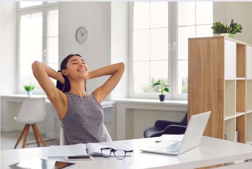 A young Asian woman leans back from her computer, done with her work in a generic room.