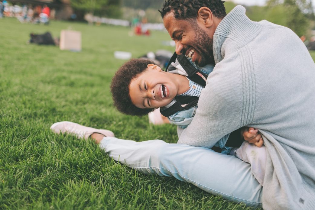 An African American Father hugging and playing with his son on the grass. 