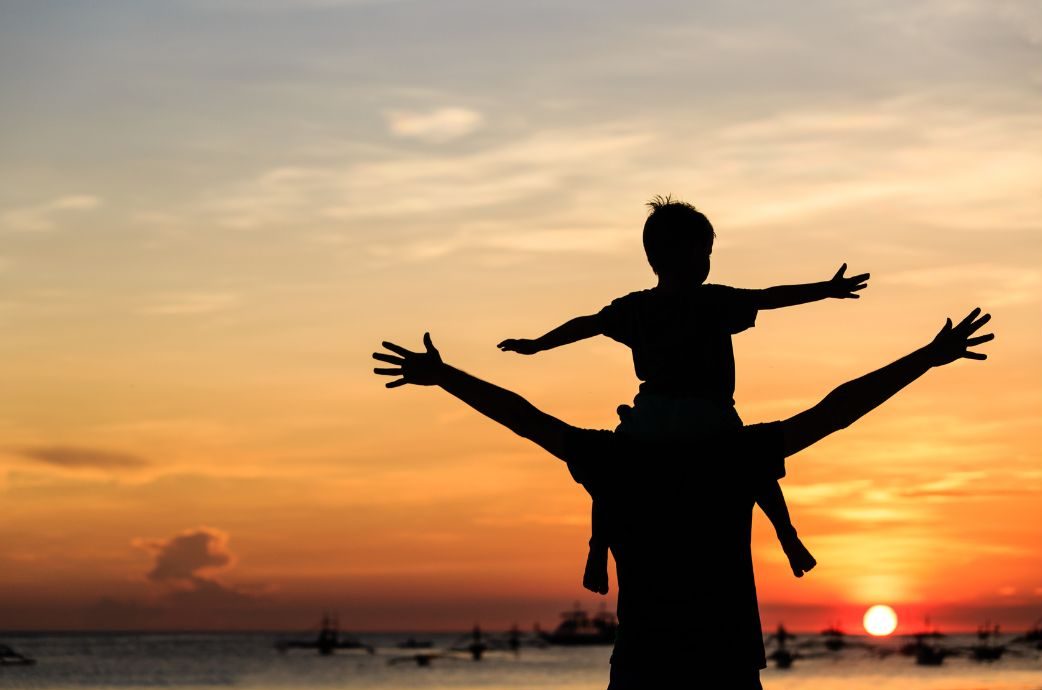 A father with his child on his shoulders with arms outstretched looking at the sunrise. 
