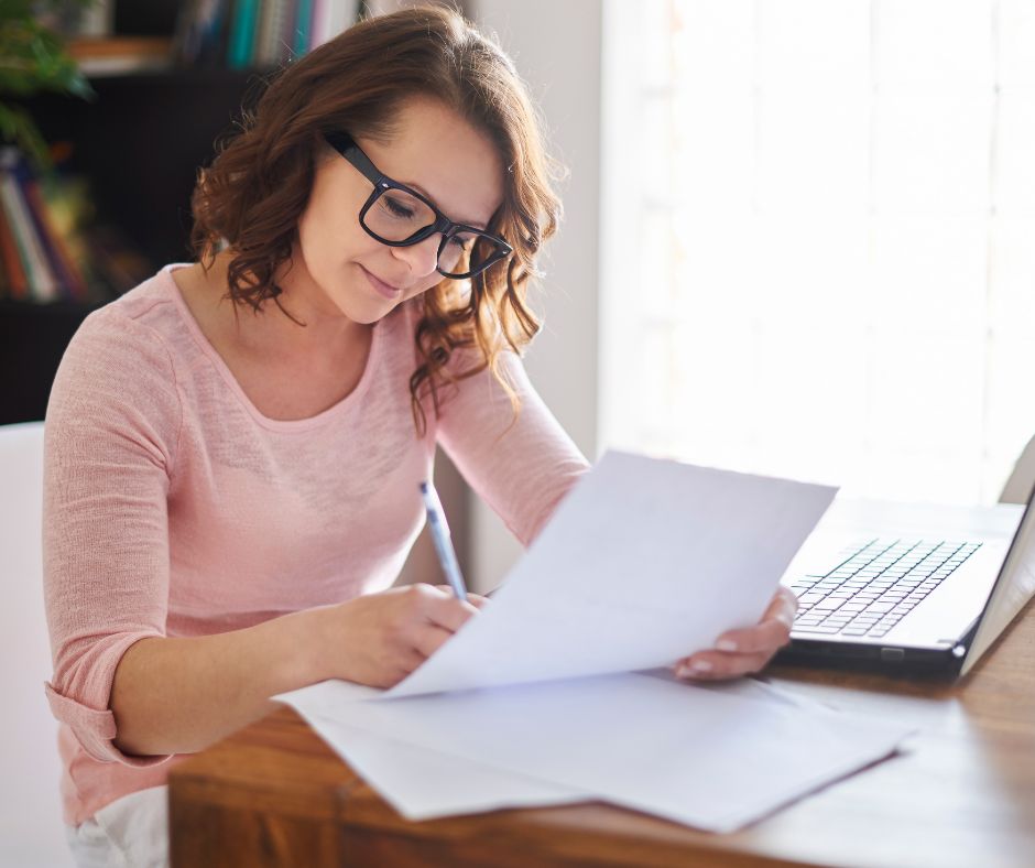 Woman, writing, papers, desk, pink shirt, glasses, window